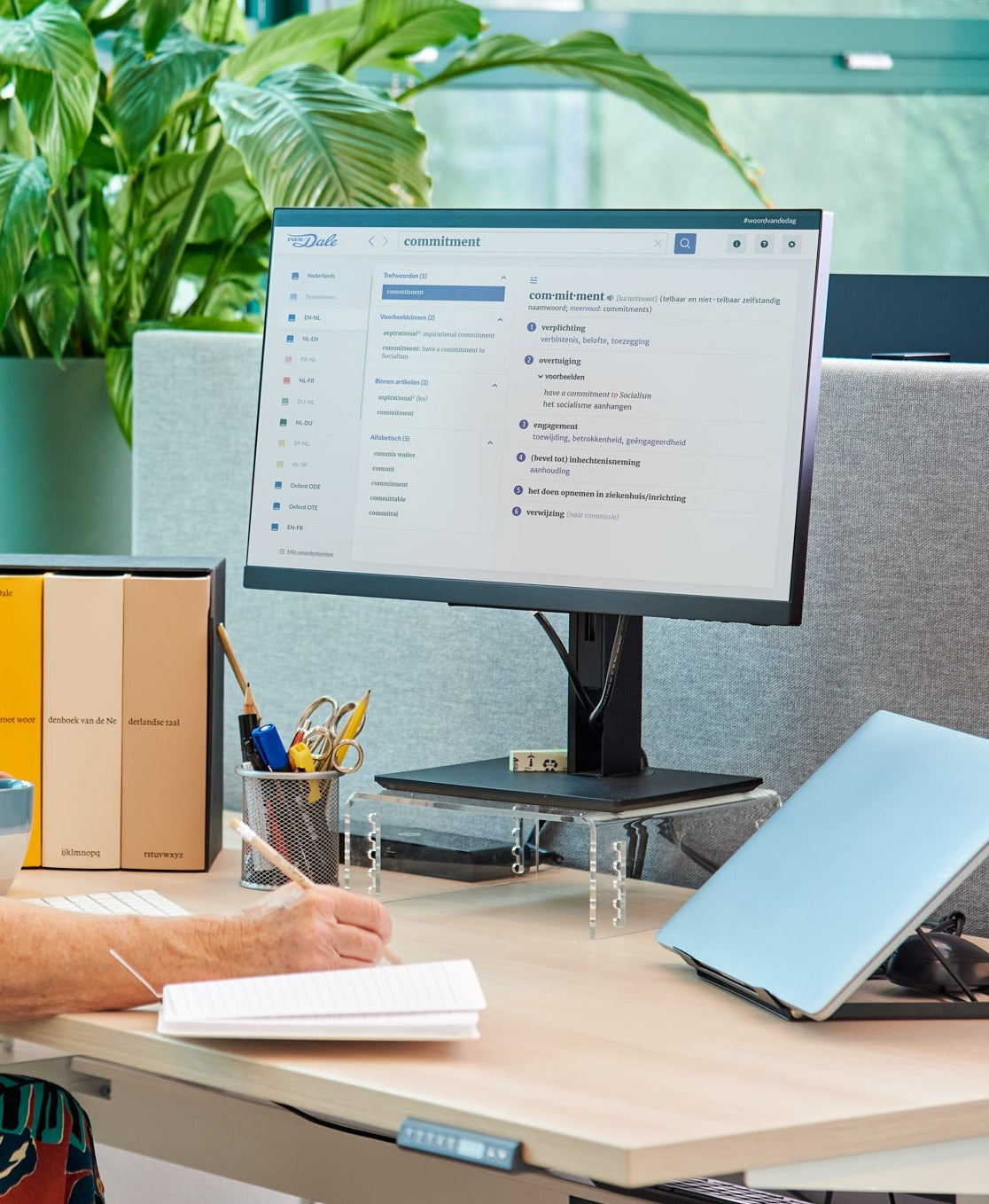 Er is een bureau te zien waar een persoon aanzit, al is van die persoon alleen de hand te zien die wat in een notitieboek opschrijft. Op het bureau staat de Dikke Van Dale en een beeldscherm met daarop Van Dale Online waarop de definitie van het woord 'commitment' te zien is