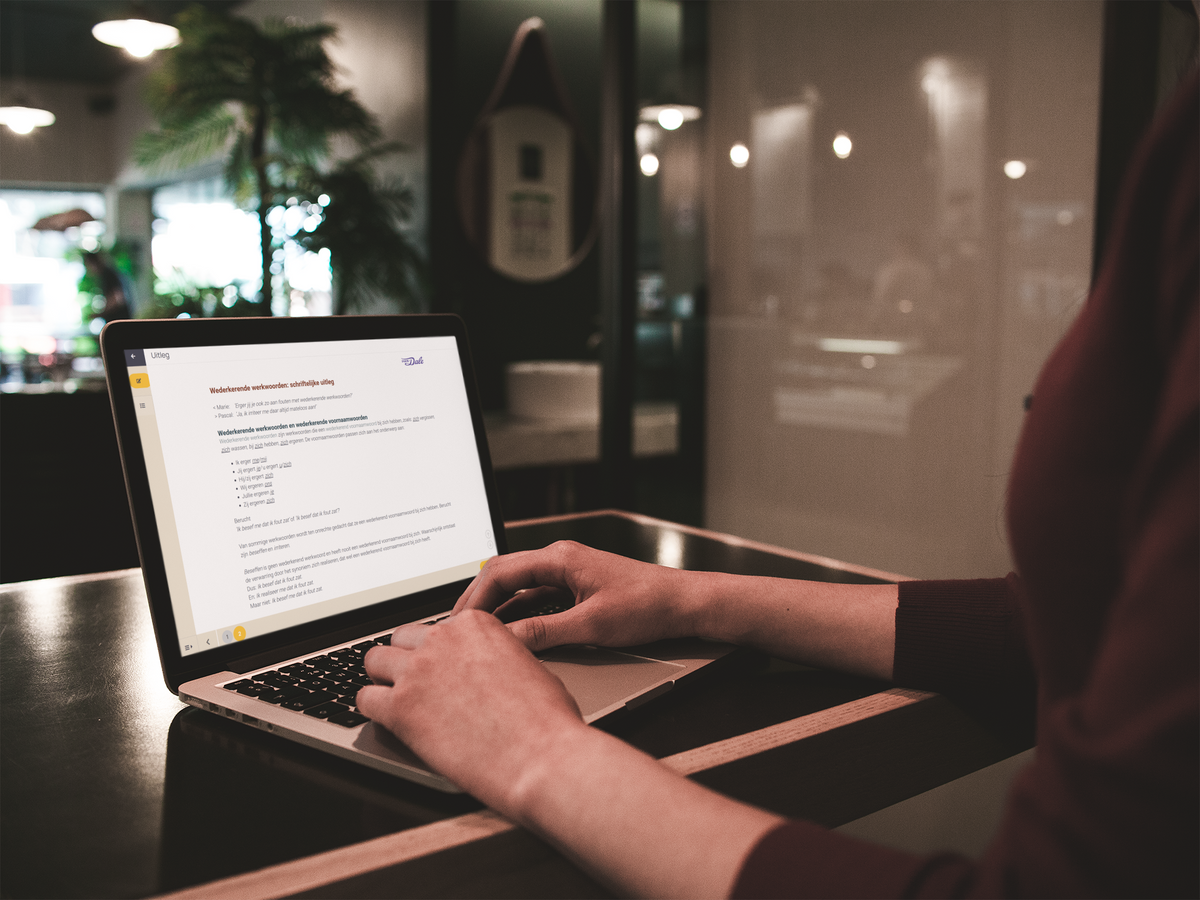 Een persoon heeft diens vingers op het toetsenbord van een laptop met daarop een schermafbeelding van een uitleg van de Van Dale E-learning werkwoordspelling