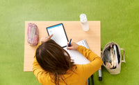 Je ziet van boven een scholier die aan tafel zit en over haar schrift gebogen is om daar wat in te schrijven. Links op tafel ligt een etui en recht ligt een tablet met daarop Van Dale Online op school. De scholier wijst met haar linkerhand naar het tabletscherm