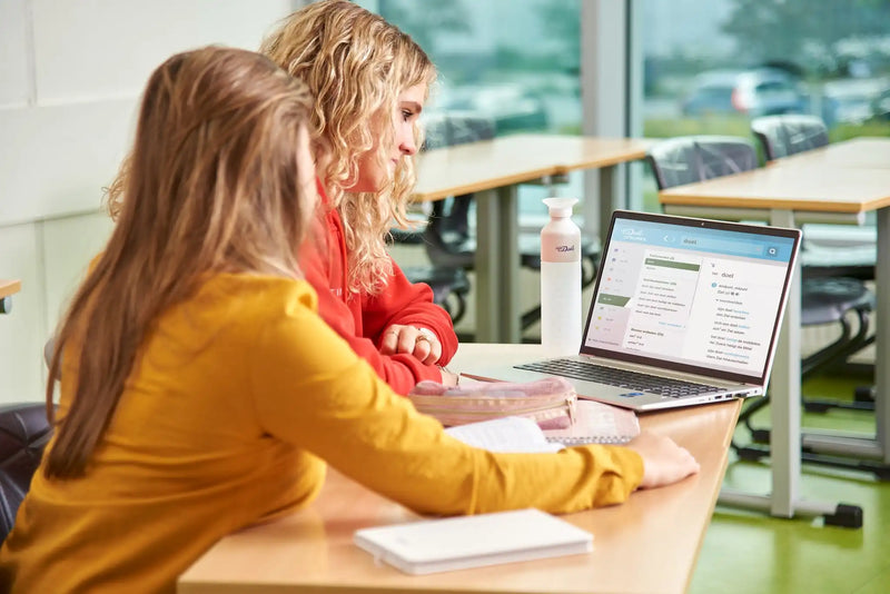 Twee scholieren zitten naast elkaar achter schooltafels en kijken naar een laptopscherm waarop Van Dale Online op school te zien is.  